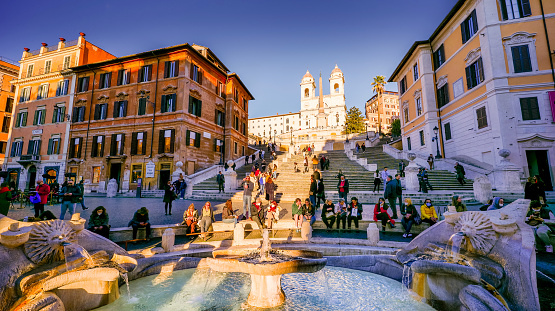 Rome, Italy, February 10 -- A warm ray of sun at sunset illuminates the Fontana della Barcaccia near the monumental stairway of Trinità dei Monti, or Spanish Steps, in the baroque heart of Rome. This iconic fountain in Rome was finished in 1629 by the architect and sculptor Pietro Bernini, father of the more famous Gian Lorenzo Bernini, commissioned by Pope Urban VIII. According to tradition, Gian Lorenzo himself participated in the final construction of the fountain on the death of his father in 1629. A masterpiece of the late Roman Baroque, the monumental stairway of the Trinità dei Monti (Spanish Steps) was built in 1721 to connect the church of the Santissima Trinità dei Monti (top in the photo) with the Piazza di Spagna (Spanish Square). Designed by the architect and urban planner Francesco De Sanctis, the Spanish Steps were inaugurated by Pope Benedict XIII on the occasion of the Jubilee of 1725. Built starting from 1502 in the Renaissance style, the church of the Santissima Trinità dei Monti was commissioned by Charles VIII, King of France, to give a stable accommodation to the religious Order of the Minims. In 1570 the façade was rebuilt based on a design by the architects Giacomo Della Porta and Carlo Maderno, with the construction of the two characteristic bell towers. In 1980 the historic center of Rome was declared a World Heritage Site by Unesco. Super wide angle image in 16:9 and high definition format.