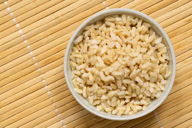 Brown cooked rice in a white ceramic bowl isolated on bamboo matt.Top view. Space for text.