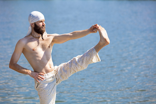 Doing yoga exercises, poses. One young bearded man meditating yoga on river berth at sunrise, outdoors. Concept of mental health, relaxation, harmony with nature. Wellness and wellbeing