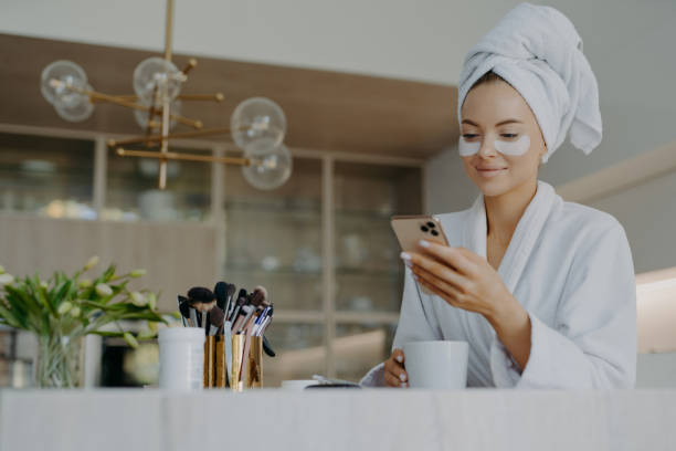 foto de atractiva mujer joven usa toalla de baño en la cabeza y bata blanca suave sostiene el teléfono móvil disfruta de la comunicación en línea aplica parches hidratantes debajo de los ojos bebidas bebidas bebidas calientes - bathrobe women cup towel fotografías e imágenes de stock