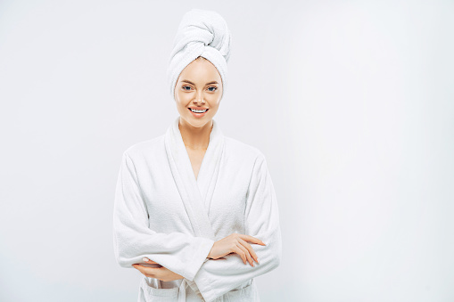 Beautiful smiling young woman has soft healthy skin after taking shower, wears bath robe and towel wrapped on head, enjoys spare time at home, isolated over white background. Wellness concept.