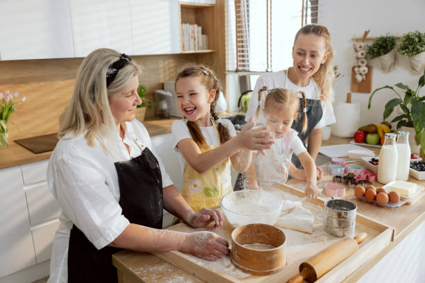 istock Elder daughter clapping hands with flour over sieve laughing having fun. Younger daughter pouring flour on sieve helping. Granny and young mom smiling. Women in the kitchen cooking 1415552388