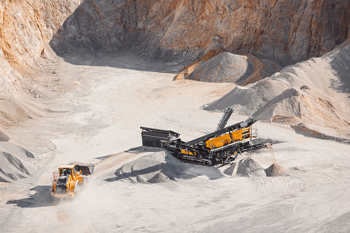 Heavy machinery in the quarry, excavator and small dump track working in the quarry. Piles of gravel all around, machines moving the gravel. Dusty roads in the quarry.