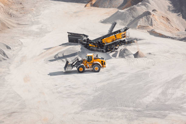 Heavy machinery in the quarry, excavator and small dump track working in the quarry. Piles of gravel all around, machines moving the gravel. Dusty roads in the quarry.