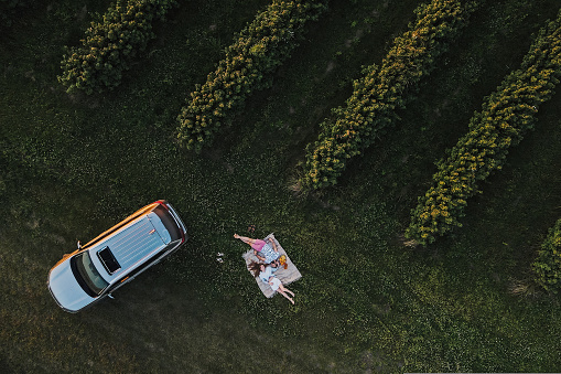 Aerial top-down shot of couple enjoying picnic on road trip in the beautiful fields, man and woman lying on blanket near the SUV car at sunset