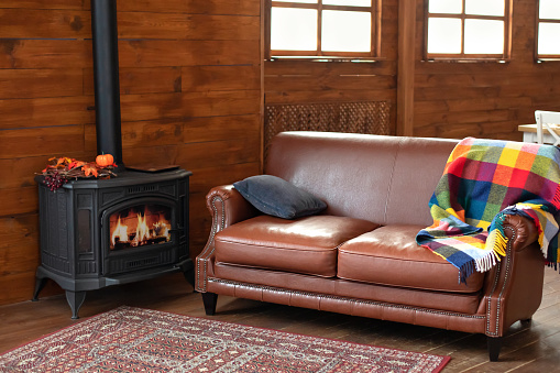 Interior cozy living room with wood burning stove inside, comfortable sofa, rustic wooden walls and carpet on floor. Black modern Cast iron wood stove at home. Fragment of interior of country house.