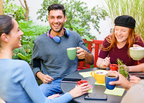 High angle of cheerful young men and women drinking coffee and laughing at joke while sitting around table during coffee break in garden