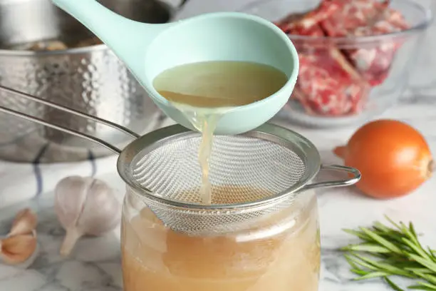 Straining delicious broth through sieve on white marble table, closeup