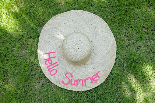 Close-up shot of a straw hat with a hello summer embroidery text, laying on the green grass