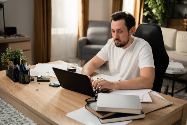 istock Middle-aged man with beard intently typing text on laptop keyboard. He loves his job, so he is responsible. On the table is cup of coffee, phone, books, calculator and stationery 1415565705