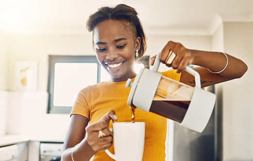Beautiful, happy and relaxed woman making coffee and pouring hot beverage in cup for morning home kitchen routine. Young smiling lady preparing or enjoying warm mug of tea, beverage or comfort drink