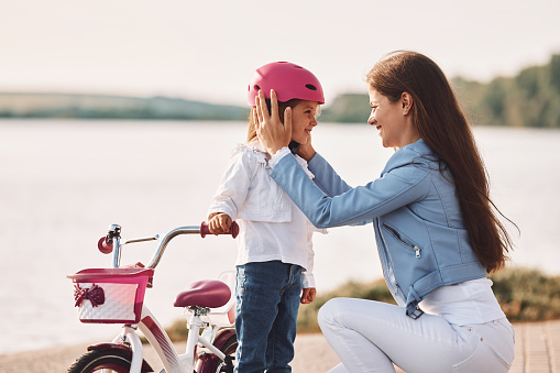 In pink colored protective helmet. Mother with her young daughter is with bicycle outdoors together.