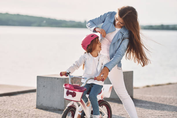a mulher está ajudando a garotinha. mãe com sua filha mais nova está com bicicleta ao ar livre juntos - pink pants summer two people - fotografias e filmes do acervo