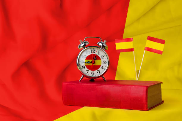 istock Time to learn Spanish. A clock on a book with two Spanish flags on a red and yellow background. Studying one of the most spoken languages in the world 1415571491