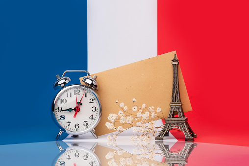 Clock and miniature of the Eiffel Tower together with an envelope to write messages with the French flag in the background. Time to learn French or travel to Paris