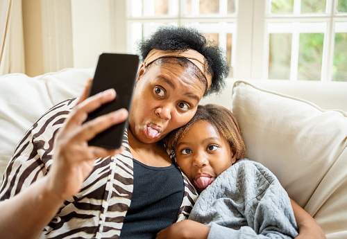 Mother and daughter sitting on sofa at home and sticking out their tongue while taking selfie with mobile phone