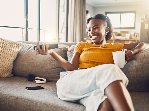 Relaxed carefree and smiling young female college student watching tv at home on her living room sofa. Happy, casual and comfortable woman sitting on her couch and enjoying her leisure free time