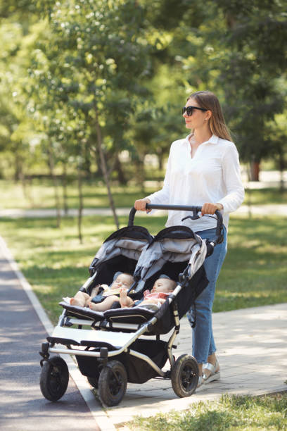 eine junge, schöne gelegenheitsmutter mit zwei zwillingen in einem doppelkinderwagen geht an einem sonnigen tag im park spazieren - sibling baby three people baby girls stock-fotos und bilder