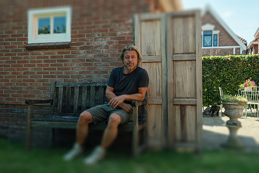 Blond man in black t-shirt, green shorts and white sneakers sits on a bench in backyard.