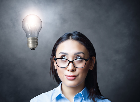 Pensive young woman with eyeglasses looking up to lightbulb light on concrete wall background. Creative thinking concept.