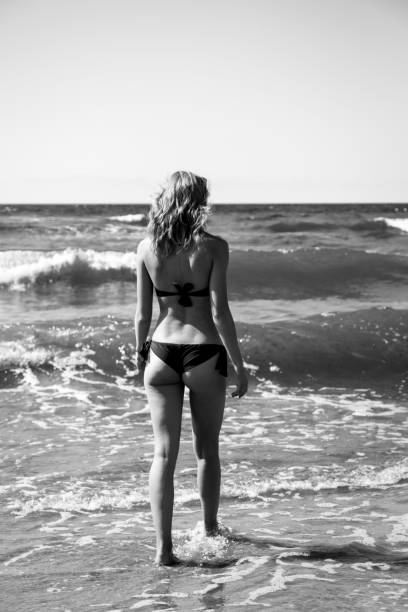 single happy young woman in swimsuit walking at sea surf with splashes, monochrome