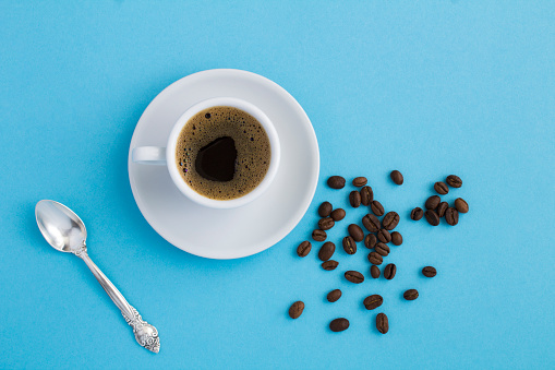 Top view of black coffee in the white cup and coffee beans on the blue background. Close-up. Copy space.