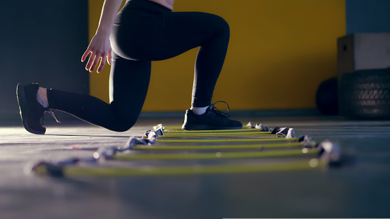 close-up, female legs in black leggings and sneakers. woman performs exercises with an athlete's foot ladder, jumps , training on agility ladder on floor, in gym. Fitness class cardio workout .