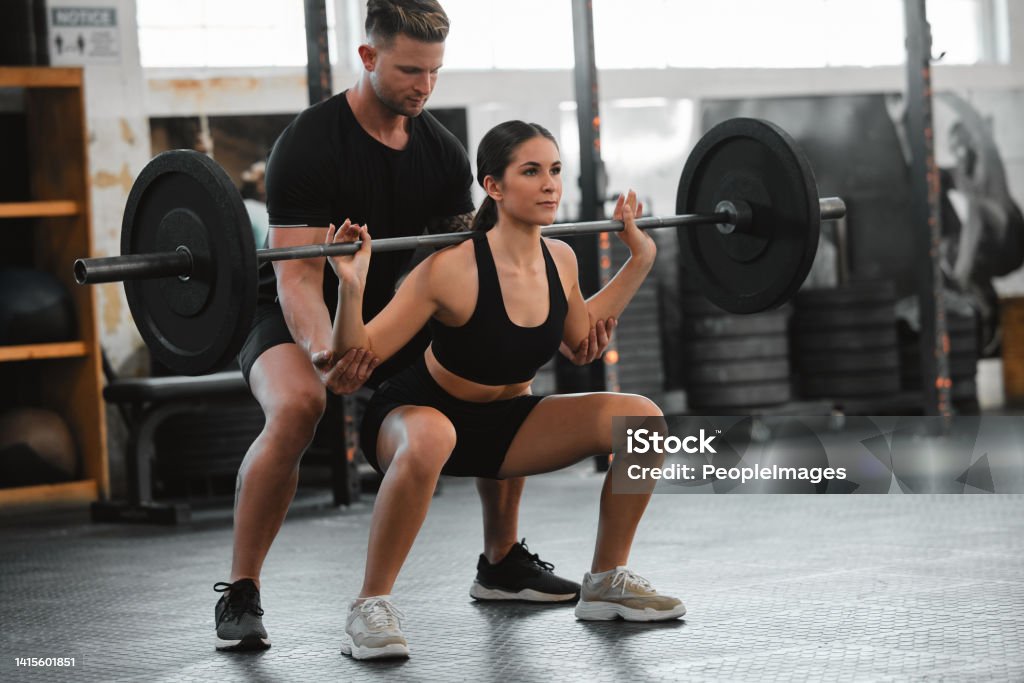 Healthy, fit and active woman exercising, weight training and squatting with her fitness and personal trainer at the gym. Athletic young female athlete lifting a barbell with her bodybuilder coach People Stock Photo