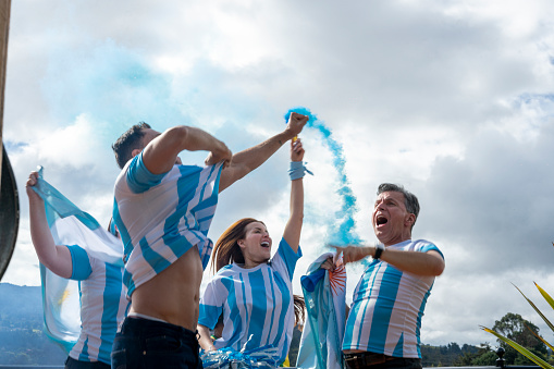 fans of the Argentinian soccer team celebrate the victory of their soccer team outdoors with blue smoke flares, flags and lots of euphoria