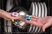 Close-up of young woman and man hands holding two colored capsule for the dishwasher. In the background, out of focus, is a dishwasher with clean dishes. Small depth of field.