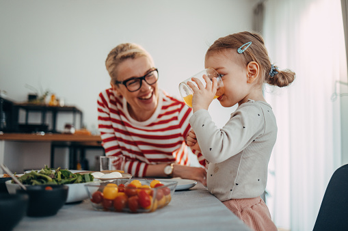 Cute young girl, standing next to the table in the kitchen, drinking a glass of orange juice in the morning. Her mom is looking at her and smiling. There is food on the table.