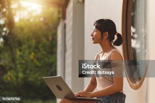 istock Young attractive Asian freelancer woman sitting on the bench in front of the window house outside working on a laptop computer during summer vacation. Happy Asian teenager student study online 1415614746