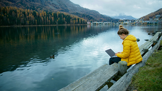 Young woman in bright yellow jacket work on side of lake on her laptop. Female freelancer work remotely during vacation. Cinematic modern millennial work style concept