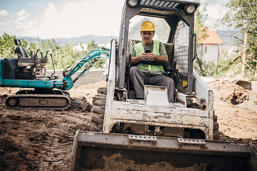 Mature construction site worker sitting in a small excavator and using smart phone at the construction site.