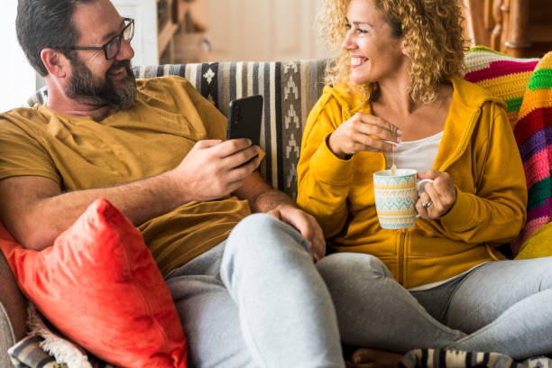 istock Smiling Man Holding Mobile Phone Sitting By Woman With Cup On Sofa 1415625909