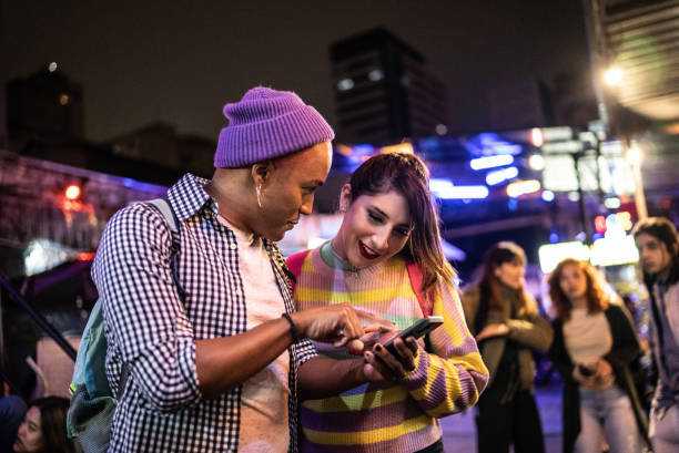 Young couple (or friends) using mobile phone at festival by the night - including a transgender person Young couple (or friends) using mobile phone at festival by the night - including a transgender person tourist couple candid travel stock pictures, royalty-free photos & images