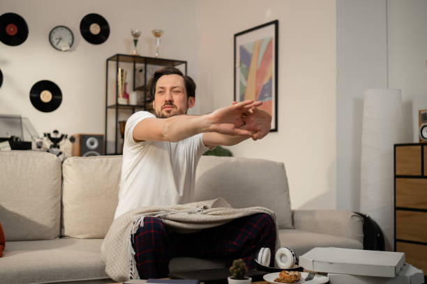 istock After hard day's work, tired man sat on couch and did some exercise. The sun shines in his face. There is pizza on table and plate of food and headphones next to it. He covered his feet with blanket 1415633756