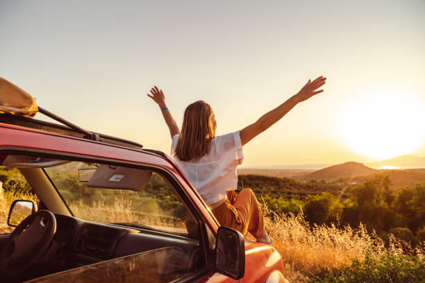 joven con los brazos en alto sentada en el coche y disfrutando de la puesta de sol - road trip fotografías e imágenes de stock