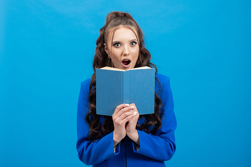 Close-up portrait of a shocked young girl hiding behind an open book, isolated on a blue wall background.