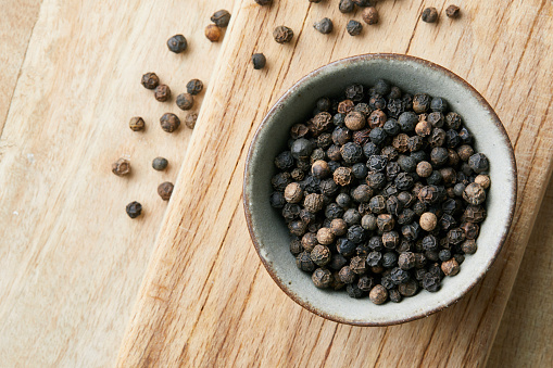 Black pepper seasoning in a small handmade ceramic bowl, on a old wooden cutting board, on a domestic kitchen table, top view with a copy space