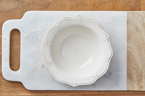 Natural white empty ceramic plate with ornaments, on a marble cutting board, on the top of a oak wooden table background