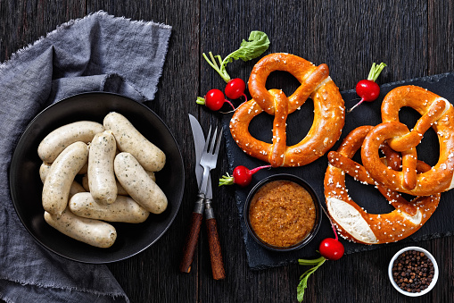 Traditional Weisswurst in black bowl, white sausages of minced veal, pork back bacon, spices and parsley, served with sweet mustard, soft pretzels, fresh red radishes, flat lay