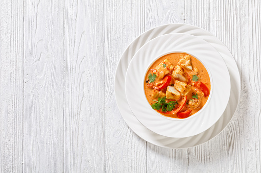 Fish Stew cooked in rich sauce of coconut milk, crushed tomatoes and sweet red pepper topped with fresh coriander in white bowl on white wood table, flat lay, free space