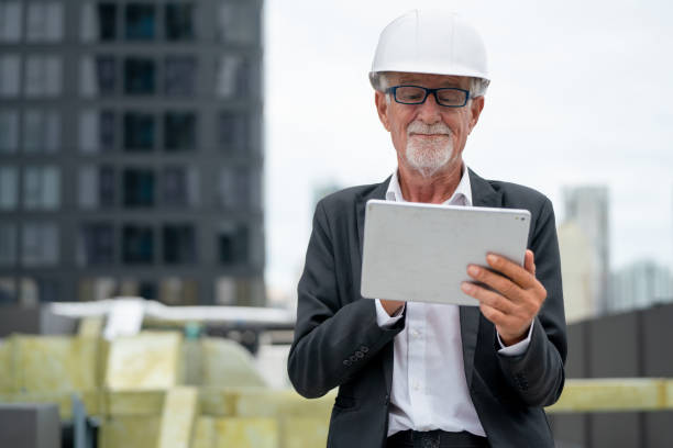 istock Senior engineer man with smiling hold tablet for his work at construction site or workplace of rooftop with other building as background. 1415650633
