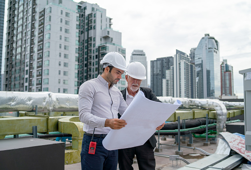 Young engineer hold drawing plan of building and discuss with senior coworker and they also stay on rooftop of construction site with cityscape as background.