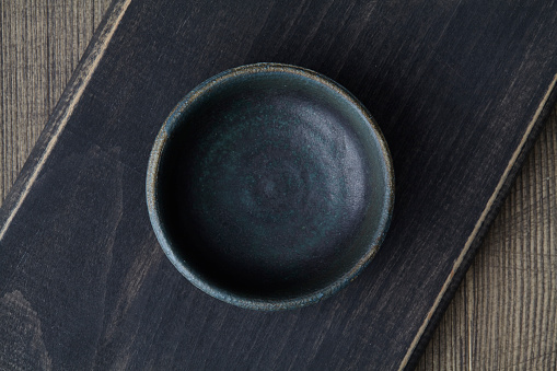 Black handmade ceramic empty bowl on a black wooden cutting board, on a rustic wooden table texture, top view with a copy space