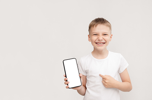 Smiling fair-haired boy in white t-shirt points his finger at the smartphone screen. copy space