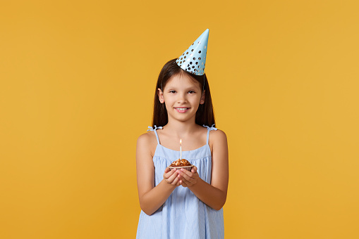 happy little birthday girl with party cone holding cupcake on yellow background