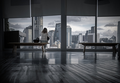 Bangkok,Thailand - Aug 13, 2022 : A calm young asia woman sitting on Modern wood bench looking through transparent wall in empty hall room with glossy floor and cityscape with skyscrapers. Rear view, Selective focus.