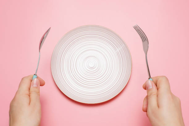 istock Female hands holding a fork and a spoon next to an empty plate on a pink background close-up top view. 1415699125